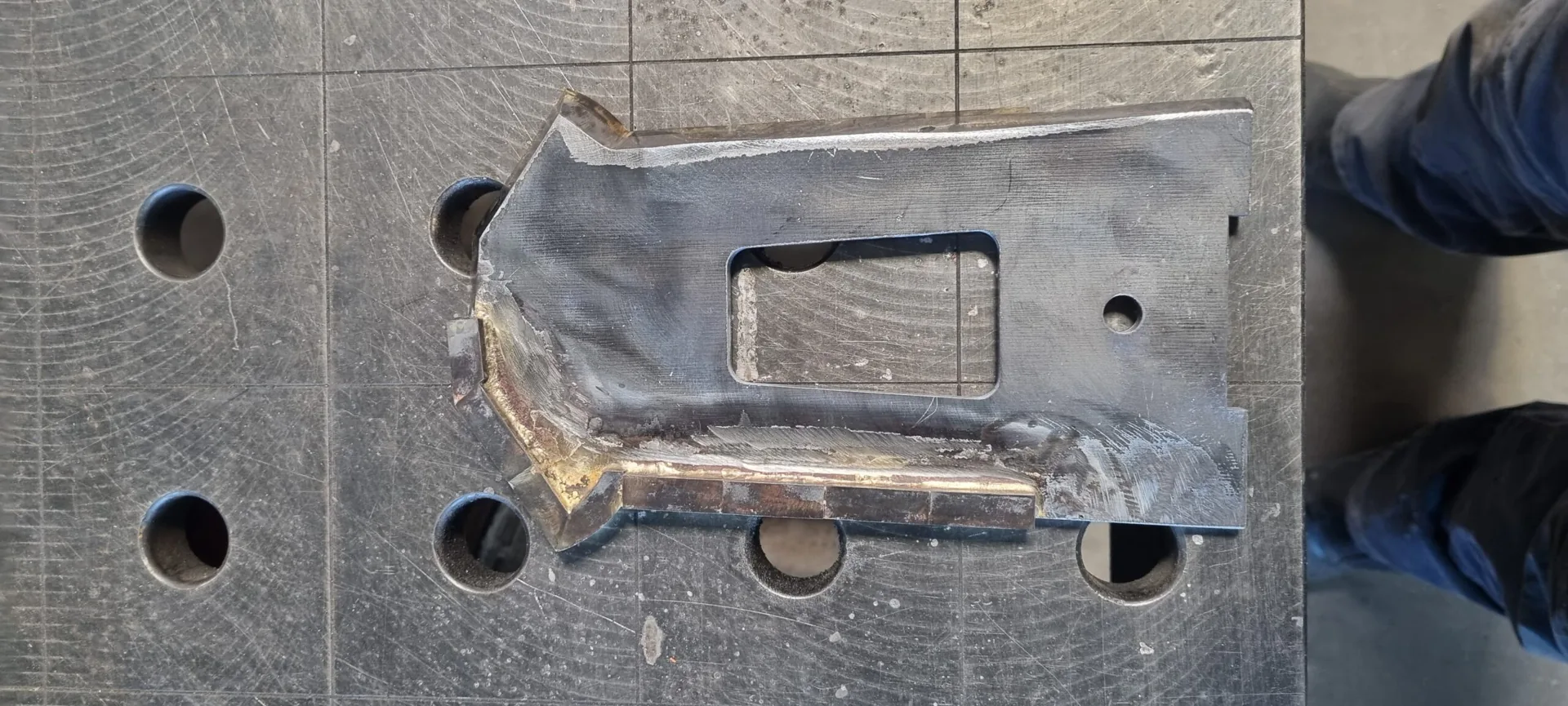 Flaches Stahlteil mit rechteckigem Durchbruch, entlang der Kante aufgelötete Hartmetall-Plättchen, von oben auf dem Schweißtisch.