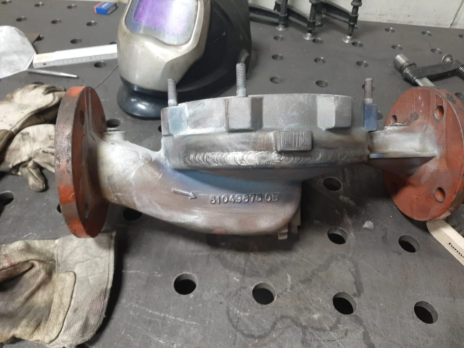 Gussgehäuse mit zwei rot lackierten Flanschen und frisch geschweißten Stellen auf dem Schweißtisch, daneben Schweißhelm und Handschuhe.